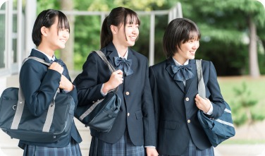 校舎の前で笑顔で並んで歩く制服姿の3人の女子生徒