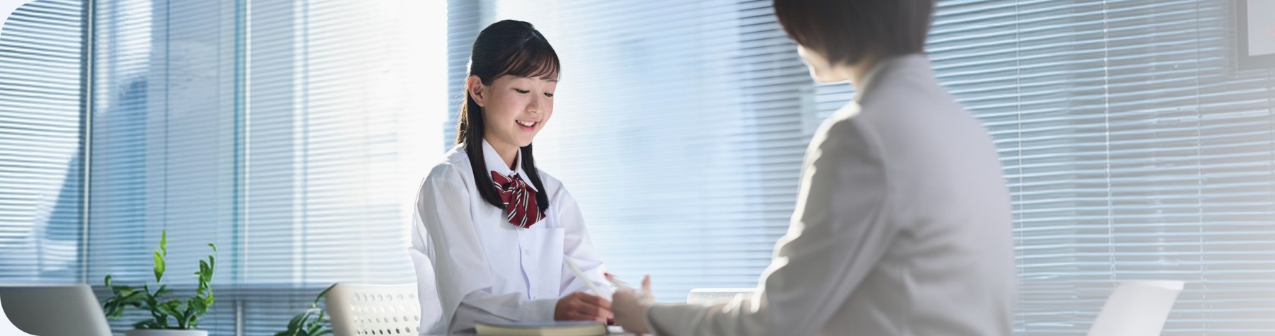 教室の机で講師と向かい合って学ぶ制服姿の女子生徒