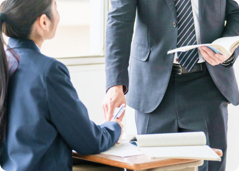 机に着いた生徒に立ったまま教材を示して説明する講師