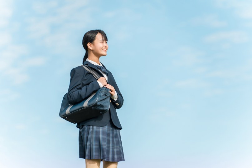 青空を背景にカバンを肩にかけて立つ制服姿の女子生徒