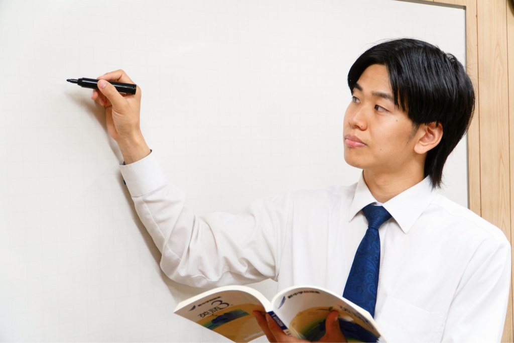 ホワイトボードにペンで書き込みながら教科書を持つ男性講師